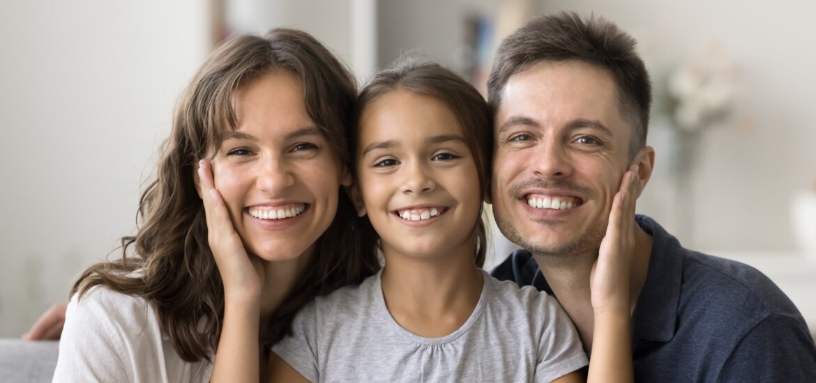 Headshot portrait cheerful smiling parents kid hug on couch at home. Adorable little girl daughter tender caress faces touch cheeks of beloved millennial mom dad