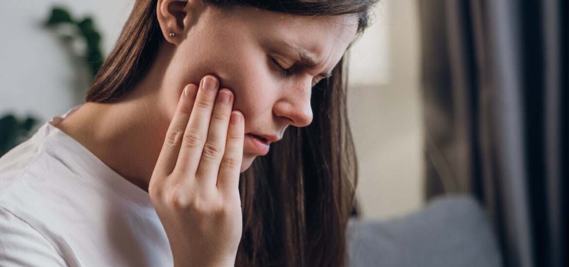 young woman suffering from TMJ related jaw pain, hand on jaw sitting on her couch