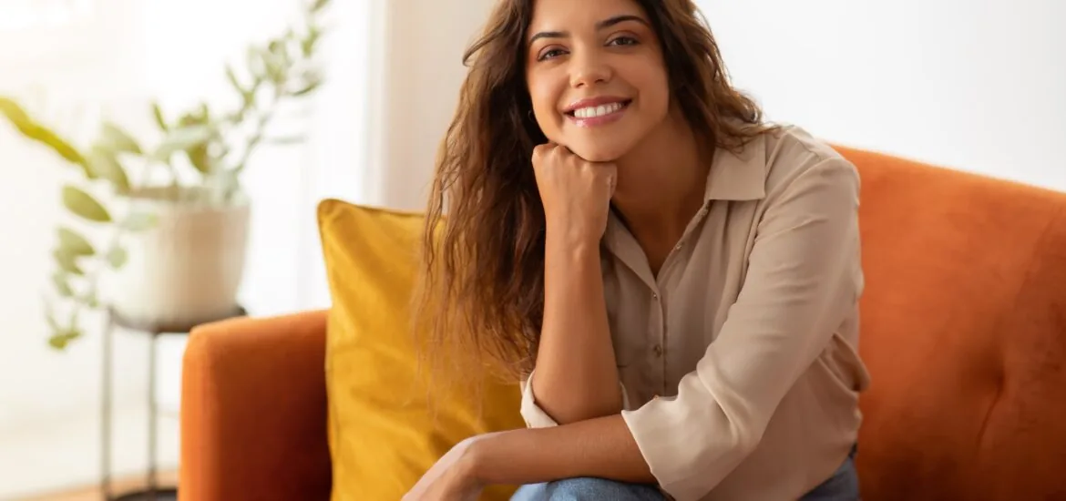 Cheerful young woman in beige shirt posing on couch at home, happy beautiful female perfect smile